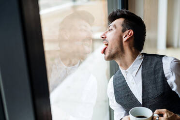 Ein junger Mann mit Kaffee steht am Fenster und streckt die Zunge heraus. - HPIF24300