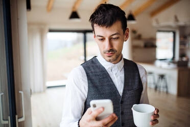 Ein junger Mann mit Kaffee und Smartphone, der zu Hause im Haus steht. - HPIF24303
