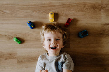 Draufsicht auf ein Kleinkind mit Spielzeugautos, die zu Hause auf dem Boden liegen. - HPIF24330