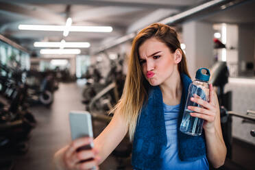 Eine junge Frau im Fitnessstudio, die mit einer Wasserflasche und einem Smartphone posiert, die Lippen spitzt und ein Selfie macht - HPIF24388