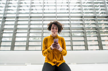 A portrait of a young student or businesswoman sitting on desk in room in a library or office, using smartphone. - HPIF24469