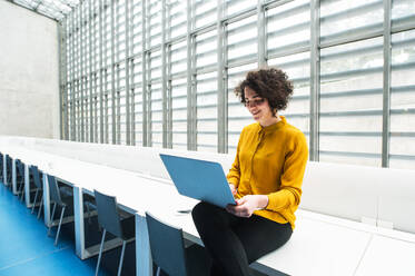 Porträt einer jungen Studentin oder Geschäftsfrau, die an einem Schreibtisch in einem Raum in einer Bibliothek oder einem Büro sitzt und einen Laptop benutzt. - HPIF24474