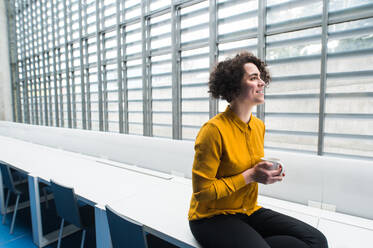 A portrait of a young student or businesswoman sitting on desk in room in a library or office, holding coffee. - HPIF24476