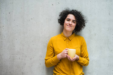 Ein Porträt einer jungen, fröhlichen Geschäftsfrau, die in einem Büro steht und eine Tasse Kaffee in der Hand hält. - HPIF24495