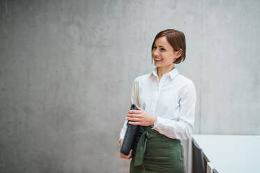 A portrait of young cheerful businesswoman standing in office, holding laptop in a bag. - HPIF24501