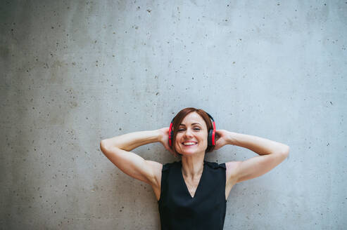 Ein Porträt einer jungen Geschäftsfrau mit Kopfhörern, die in einem Büro an einer Betonwand steht und mit geschlossenen Augen Musik hört. - HPIF24520