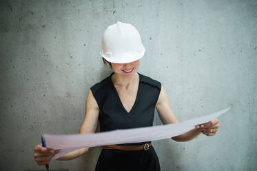 Eine junge Geschäftsfrau oder Architektin mit weißem Helm steht in einem Büro und betrachtet Baupläne. - HPIF24529