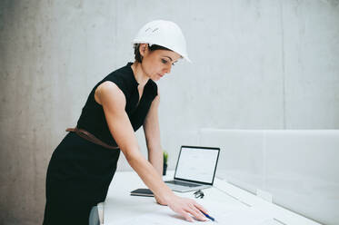 Eine junge Geschäftsfrau oder Architektin mit Helm und Laptop steht am Schreibtisch im Büro. - HPIF24530