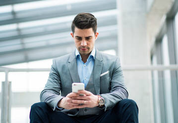 Ein Porträt eines glücklichen jungen Geschäftsmannes mit Smartphone, der im Korridor vor seinem Büro sitzt. - HPIF24539