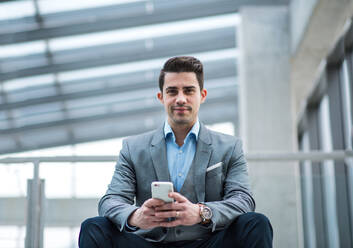 Ein Porträt eines glücklichen jungen Geschäftsmannes mit Smartphone, der im Korridor vor seinem Büro sitzt. - HPIF24540