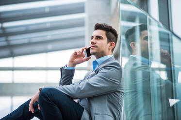 A portrait of happy young businessman with smartphone sitting in corridor outside office, making a phone call. - HPIF24541