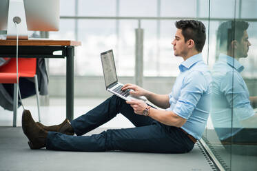 A portrait of young businessman with laptop sitting on the floor in office, working. - HPIF24545