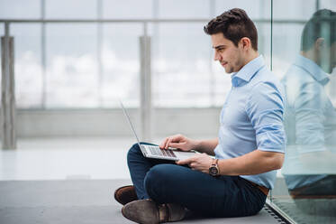 Ein Porträt eines jungen Geschäftsmannes mit Laptop, der auf dem Boden im Korridor vor seinem Büro sitzt. - HPIF24546