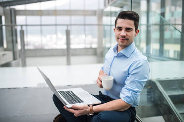 A portrait of young businessman with laptop and coffee sitting on the floor in corridor outside office. - HPIF24548