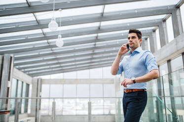 A low-angle view of young serious businessman with smartphone in corridor outside office, making a phone call. - HPIF24553