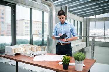 Junger Geschäftsmann oder Architekt mit Modell eines Hauses, der am Schreibtisch im Büro steht und sich bei der Arbeit Notizen macht. - HPIF24560