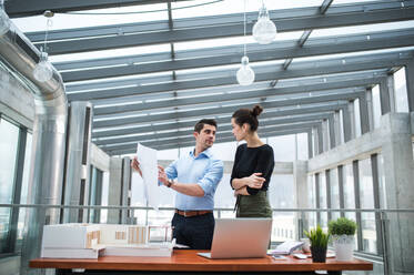 Two young male and female architects with laptop, blueprints and model of a house standing in office, talking. - HPIF24568