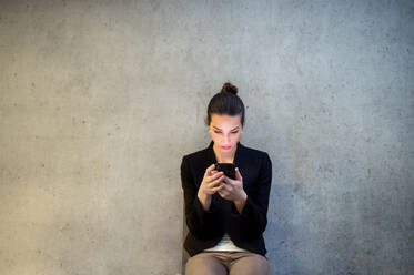 Frontansicht einer glücklichen jungen Geschäftsfrau mit Smartphone, die in einem Büro an einer Betonwand sitzt. - HPIF24598