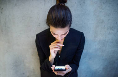 Eine Draufsicht auf eine glückliche junge Geschäftsfrau mit Smartphone, die an einer Betonwand in einem Büro steht. - HPIF24600