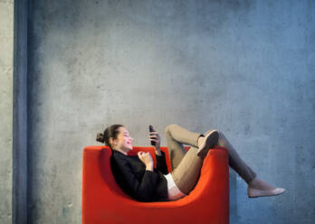 A young businesswoman with smartphone sitting on red armchair in office, a gray concrete wall in the background. - HPIF24601