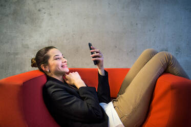 A young businesswoman with smartphone sitting on red armchair in office, a gray concrete wall in the background. - HPIF24602