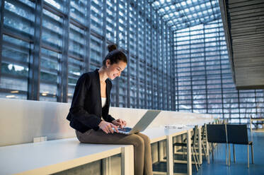 A portrait of a young student or businesswoman sitting on desk in room in a library or office, using laptop. - HPIF24608