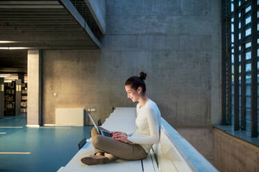 A portrait of a young student or businesswoman sitting on desk in room in a library or office, using laptop. - HPIF24616