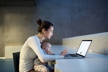 Eine Seitenansicht einer jungen Studentin, Mutter oder Geschäftsfrau, die mit einem Baby in einem Raum in einer Bibliothek oder einem Büro am Schreibtisch sitzt und einen Laptop benutzt. - HPIF24618