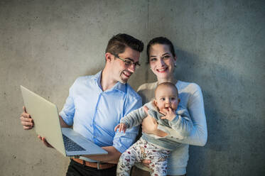 A young businessman with wife and baby daughter standing together in office, using laptop. - HPIF24621