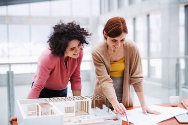 Zwei junge Architektinnen mit einem Hausmodell stehen im Büro, arbeiten und unterhalten sich. - HPIF24671