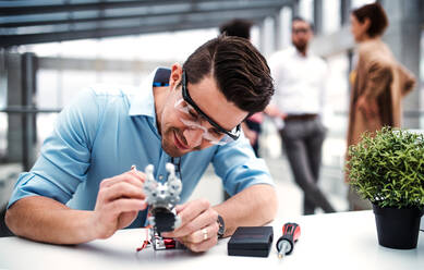 A young businessman or scientist with robotic hand and safety glasses standing in office, working. - HPIF24677