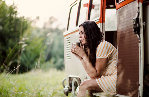 Ein fröhliches junges Mädchen sitzt im Kofferraum eines Autos auf einem Roadtrip durch die Landschaft und trinkt Kaffee. - HPIF24716