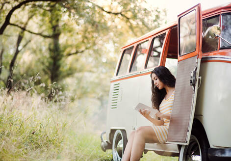 Ein fröhliches junges Mädchen mit einem Buch am Auto auf einem Roadtrip durch die Landschaft, lesend. Kopierraum. - HPIF24717