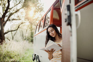 Ein fröhliches junges Mädchen mit einem Buch am Auto auf einem Roadtrip durch die Landschaft, lesend. Kopierraum. - HPIF24719