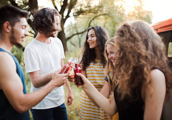 Eine Gruppe junger Freunde steht mit Getränken im Freien auf einem Roadtrip durch die Landschaft und stößt mit Flaschen an. - HPIF24743