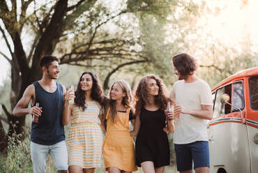 A group of young friends walking by a retro minivan on a roadtrip through countryside, holding bottles. - HPIF24744