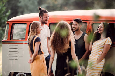 Eine Gruppe junger Freunde steht lachend neben einem Retro-Minivan auf einem Roadtrip durch die Landschaft. - HPIF24748