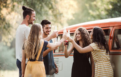 Eine Gruppe junger Freunde steht mit Getränken im Freien auf einem Roadtrip durch die Landschaft und stößt mit Flaschen an. - HPIF24750