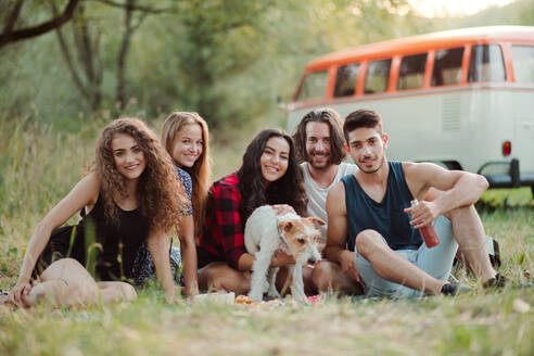 Eine Gruppe junger Freunde mit einem Hund sitzt im Gras vor einem Retro-Minivan auf einem Roadtrip durch die Landschaft. - HPIF24764