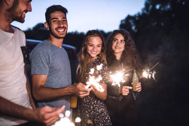 Eine Gruppe junger Freunde mit Wunderkerzen steht in der Abenddämmerung auf einem Roadtrip durch die Landschaft. - HPIF24768