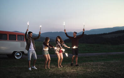 Eine Gruppe junger Freunde mit Wunderkerzen steht in der Abenddämmerung auf einem Roadtrip durch die Landschaft. - HPIF24777