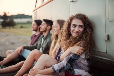 Eine Gruppe junger Freunde auf einem Roadtrip durch die Landschaft, die neben einem Retro-Minivan sitzen. - HPIF24779