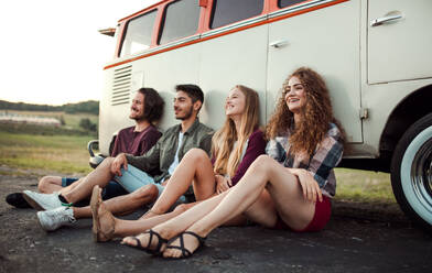 Eine Gruppe junger Freunde auf einem Roadtrip durch die Landschaft, die neben einem Retro-Minivan sitzen. - HPIF24780