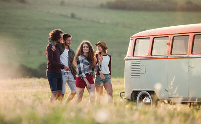 A group of young friends walking in front of a retro minivan on a roadtrip through countryside, walking arm in arm. - HPIF24794