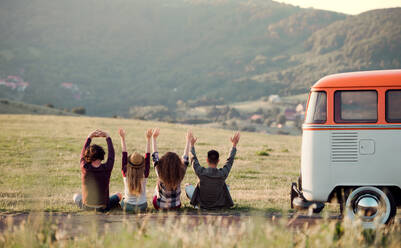 Rückansicht einer Gruppe junger Freunde, die auf einem Roadtrip durch die Landschaft im Gras sitzen und die Arme ausstrecken. - HPIF24795