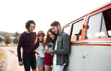 Eine Gruppe fröhlicher junger Freunde mit einem Retro-Minivan auf einem Roadtrip durch die Landschaft, mit Smartphone. - HPIF24804