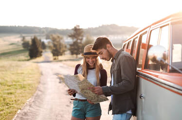 Ein junges Paar auf einem Roadtrip durch die Landschaft, das neben einem Retro-Minivan steht und auf eine Landkarte schaut. - HPIF24812