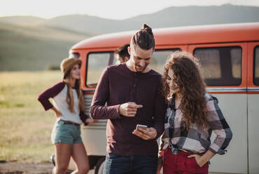 Ein junges Paar auf einem Roadtrip durch die Landschaft, das an einem Minivan steht und eine Karte auf dem Smartphone benutzt. - HPIF24813