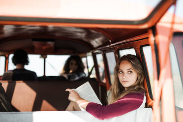 Eine Gruppe fröhlicher junger Freunde mit einem Retro-Minivan auf einem Roadtrip durch die Landschaft, ein Mädchen liest ein Buch. - HPIF24842