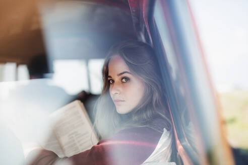 Eine Gruppe junger Freunde mit einem Retro-Minivan auf einem Roadtrip durch die Landschaft, ein Mädchen liest ein Buch, aufgenommen durch Glas. - HPIF24843